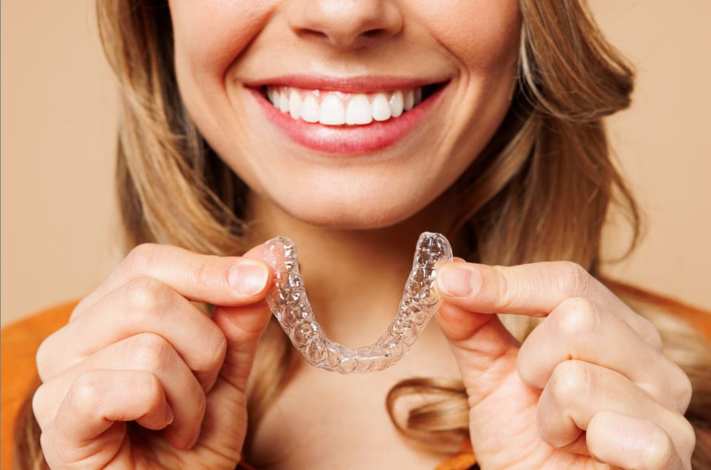 Woman Smiling And Holding A Clear Invisalign Aligner Close To Her Teeth Woman Smiling And Holding A Clear Invisalign Aligner Close To Her Teeth